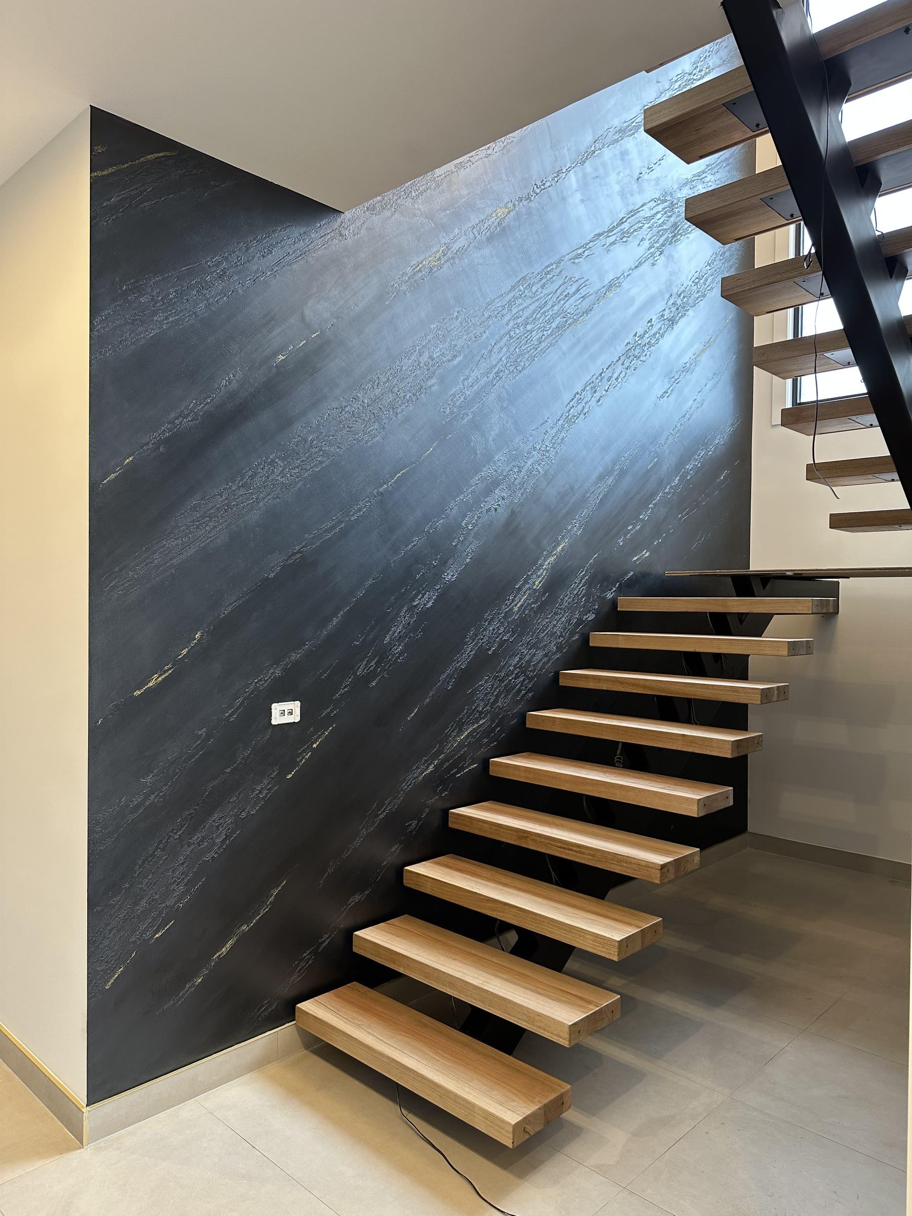 A full-height wall in black split-stone plaster with pale grey veining running diagonally through it, floating oak stair treads on a black steel stringer rising in front of it above pale tiles